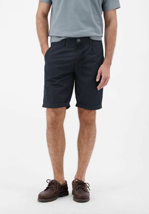 Mann trägt dunkelblaue Shorts mit umgeschlagenem Saum, hellgraues Poloshirt und braune Leder-Schnürschuhe, steht mit einer Hand in der Hosentasche.