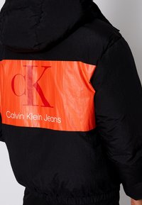 Calvin Klein Jeans pufferjacka med svart yttre och en livlig orange panel med vit logotext. Huva och strukturerad yta.