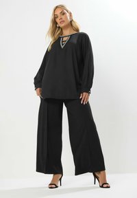 Maglietta nera a maniche lunghe con colletto decorato di gioielli e dettagli a taglio, abbinata a pantaloni neri a gamba larga e sandali neri con tacco.