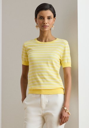 Frau trägt ein gelb-weiß gestreiftes kurzärmeliges Oberteil mit weißen Hosen, Muschel-Ohrringe und ein passendes Muschelarmband, steht drinnen.