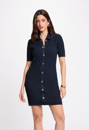 Kvinde iført en kortærmet tekstureret navyblå kjole med metalknapper foran og krave, accessoriseret med gyldne hoop-øreringe og armbånd.