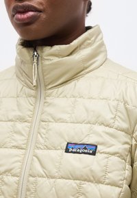 Κοντινό πλάνο ενός ατόμου που φοράει μία μπεζ, καπιτονέ μπουφάν Patagonia με ψηλό, φερμουάρ γιακά και λογότυπο στο στήθος.