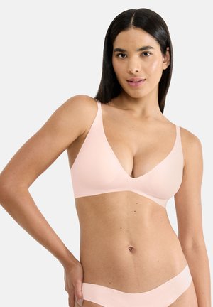 Žena se středním odstínem pleti a rovnými tmavými vlasy, oblečená v růžovém bezdrátovém podprsence a odpovídajícím spodním prádle, pózující s rukou na boku.