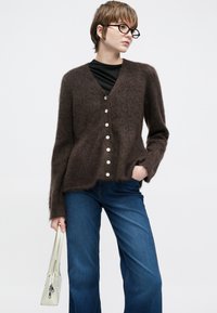 Καφέ, αφράτη ζακέτα με V-λαιμόκοψη, με έξι μεγάλα λευκά κουμπιά και μακριά μανίκια. Συνδυασμένη με τζιν παντελόνι φαρδύ στα πόδια και ελαφριά τσάντα.