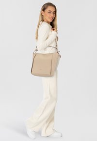Beige Leder-Shopper mit einer strukturierten Form, ausgestattet mit einem oberen Griff und einem verstellbaren Schultergurt. Glatte Textur und dezentes Logo-Detail auf der Vorderseite.