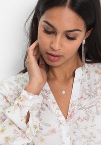 Blusa bianca con motivo floreale, collana d'argento con un pendente rotondo e orecchini lunghi d'argento; texture liscia, design delicato.