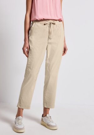 Pantaloni cargo beige, tapered, con tasche laterali, vita elasticizzata e dettaglio con cordino, abbinati a sneakers bianche e beige.