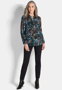 Blusa a maniche lunghe di colore verde scuro con un motivo di piume colorate, abbinata a pantaloni neri aderenti e stivali neri alla caviglia.