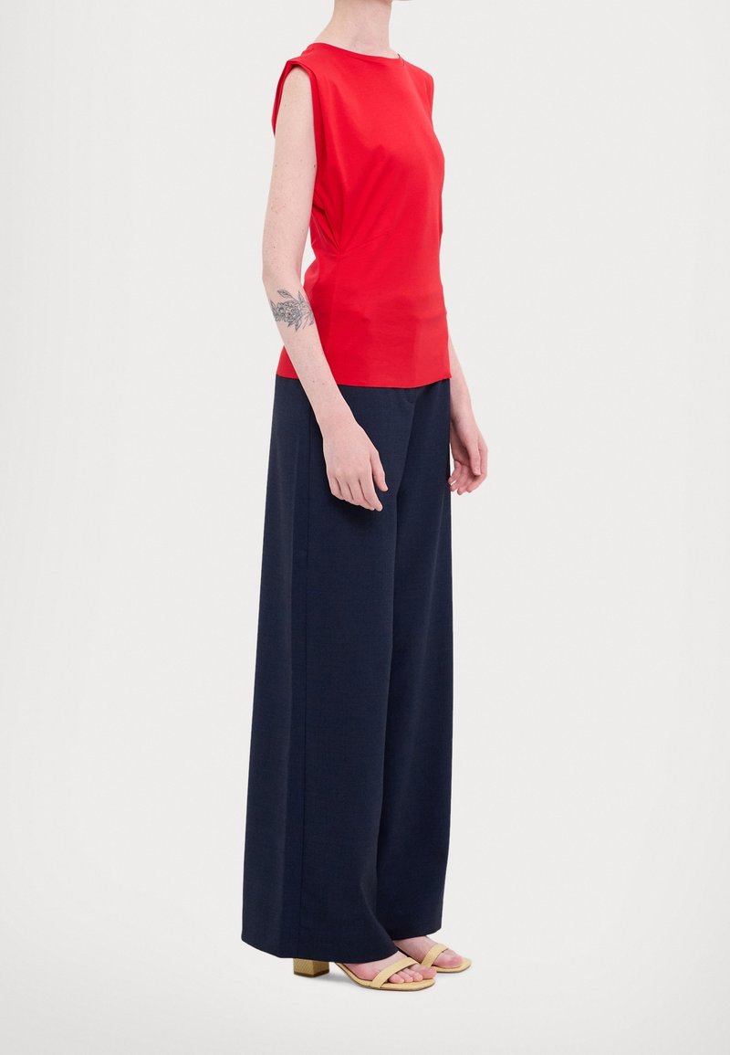 Personne portant un haut rouge sans manches, un pantalon large bleu marine, des sandales beiges à talons et un tatouage floral sur l'avant-bras gauche.