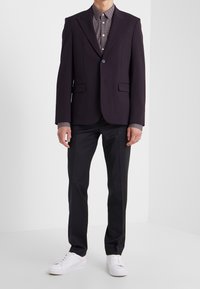 Blazer viola scuro con due tasche frontali, abbinato a una camicia chiara a pois e pantaloni scuri, completato da sneaker bianche.