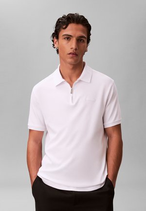 Młody mężczyzna o ciemnych kręconych włosach, ubrany w biały, krótki rękaw koszulki polo z zamkiem z przodu, stoi z rękami w kieszeniach czarnych spodni.