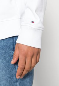 Sweatshirt blanc avec des poignets côtelés, présentant un petit logo bleu, rouge et blanc sur la manche. La main repose contre un tissu en denim.