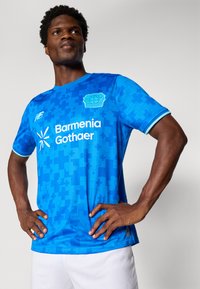 Man standing with hands on hips wearing blue Bayer Leverkusen soccer jersey and white shorts against plain background.