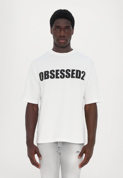 Weißes Baumwoll-T-Shirt mit kurzen Ärmeln, das auf der Vorderseite den auffälligen schwarzen Text "OBSESSED2" trägt. Getragen mit hellgrauen, zerrissenen Jeans.