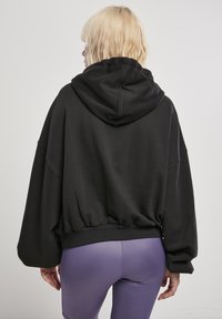 Felpa nera corta con cappuccio regolabile, maniche larghe e fondo arricciato. Il materiale appare morbido e strutturato. Indossata sopra leggings viola.