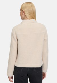 Lichtbeige textuurjasje met een kraag, elastische zoom en lange mouwen. Het materiaal lijkt zacht en heeft een visgraatpatroon.