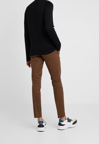 Person in braunen Slim-Fit-Hosen, schwarzem Langarmshirt und weißen Sneakers mit blauen, grünen und gelben Akzenten, steht mit den Händen in den Taschen.