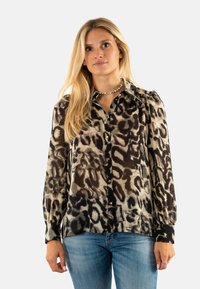 Blusa de estampado de leopardo hecha de tela ligera, con mangas largas, tapeta de botones y puños negros. Los colores incluyen beige, marrón y negro.