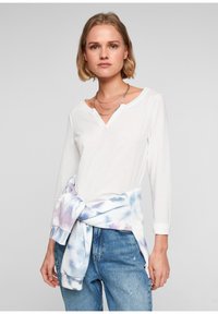 Biała koszula z długim rękawem, z dekoltem w serek, oraz bluzą w tie-dye związaną w pasie. Niebieskie dżinsy z lekkim przetarciem dopełniają stylizację.