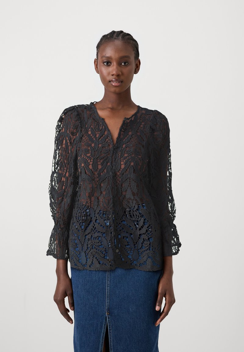 Blouse en dentelle noire avec des motifs floraux intricats, des manches longues et un décolleté en V. Présente un aspect semi-transparent et des bords festonnés.