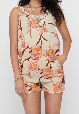 Femme portant une combinaison beige sans manches avec un imprimé floral rose et jaune, main dans la poche, accessoirisée avec des colliers argentés superposés et un bracelet.