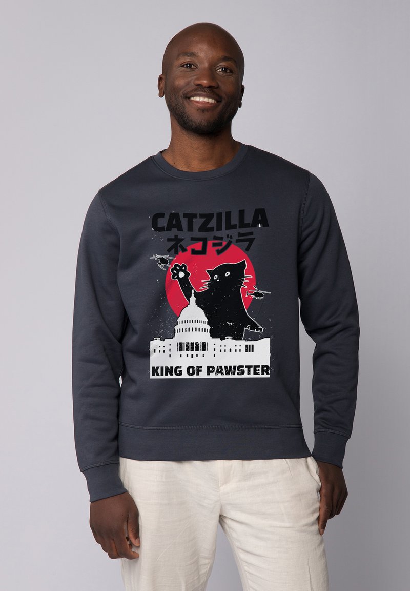 Grijze sweatshirt met grafische print van een zwarte kat tegen een rode cirkel, een witte gebouw, en de tekst "CATZILLA" en "KING OF PAWSTER."
