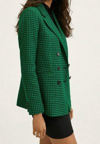 Blazer vert à double boutonnage avec motif pied-de-poule, col proéminent, boutons noirs, et coupe structurée, associé à une jupe noire.