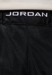 Ceinture en tissu noir avec "JORDAN" brodé en blanc. Surface texturée avec des lignes parallèles, affichant un design sportif.