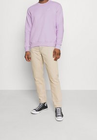 Sudadera de un suave color púrpura claro con cuello redondo y logotipo blanco, combinada con pantalones beige y zapatillas altas negras. Textura de tejido suave.
