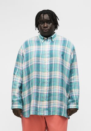 Persona negra con rastas cortas que lleva una camisa de cuadros suelta en tonos teal, blanco y púrpura abotonada, y pantalones color coral, mirando al frente.