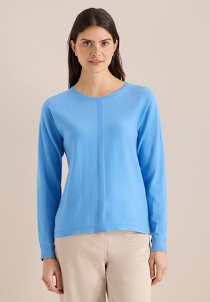 Femme aux cheveux longs et foncés portant un pull à manches longues bleu clair et un pantalon beige, debout devant un fond clair uni.
