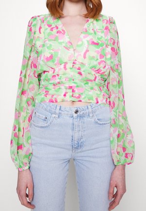 Blusa floreale verde e rosa con maniche a sbuffo lunghe e scollo a V, caratterizzata da un design avvolgente e arricciature in vita. Jeans in denim chiaro.