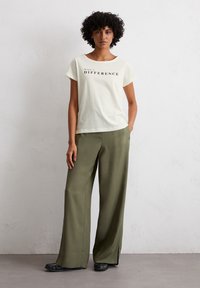T-shirt en coton blanc avec le texte "FAITES UNE DIFFÉRENCE", associé à un pantalon large olive. Des chaussures plates noires complètent le look.