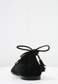 Zapato plano de ante negro con punta redonda, cierre en la parte posterior con lazo y dos borlas decorativas en el talón. Textura suave y diseño minimalista.