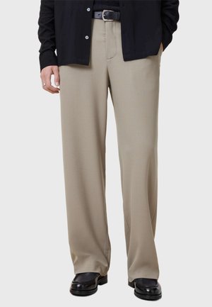 Pantalons larges beiges en tissu lisse, avec une couture plate, taille mi-haute et pli subtil, associés à des chaussures noires.