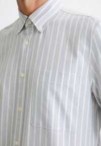 Camicia button-up grigia chiara con righe verticali bianche, colletto e tasca sul petto. Il tessuto appare liscio con una sottile texture.