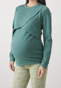 Mörkt tealfärgad långärmad gravidtröja med ett lager-på-lager design. Kombination med texturerade gröna och gula mönstrade byxor.