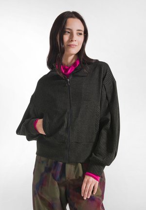 Woman with dark hair wears a black zip-up jacket with thin stripes, bright pink top underneath, and multicolored loose pants, standing against white.