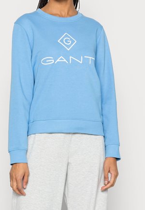 Femme portant un sweat GANT bleu clair à manches longues et un pantalon gris clair sur un fond uni.