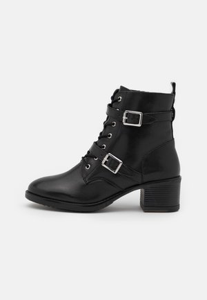 Musta nahkainen nilkkuri, jossa on paksu korko, edessä nyöritys ja kaksi säädettävää hopeista solkea valkoisella taustalla.