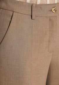 Beige op maat gemaakte broeken met voorzakken, een riemlus en een knoopsluiting die de stoftextuur en stikseldetails toont.