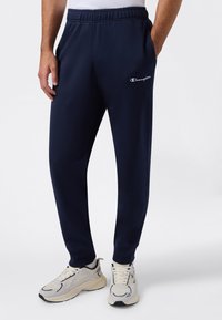 Hombre vistiendo pantalones de chándal azul marino de Champion y zapatillas deportivas blancas, de pie con las manos en los bolsillos sobre un fondo blanco.