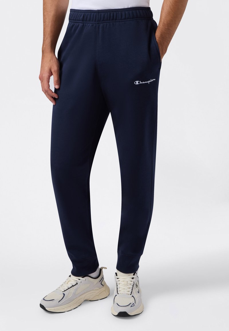 Hombre vistiendo pantalones de chándal azul marino de Champion y zapatillas deportivas blancas, de pie con las manos en los bolsillos sobre un fondo blanco.