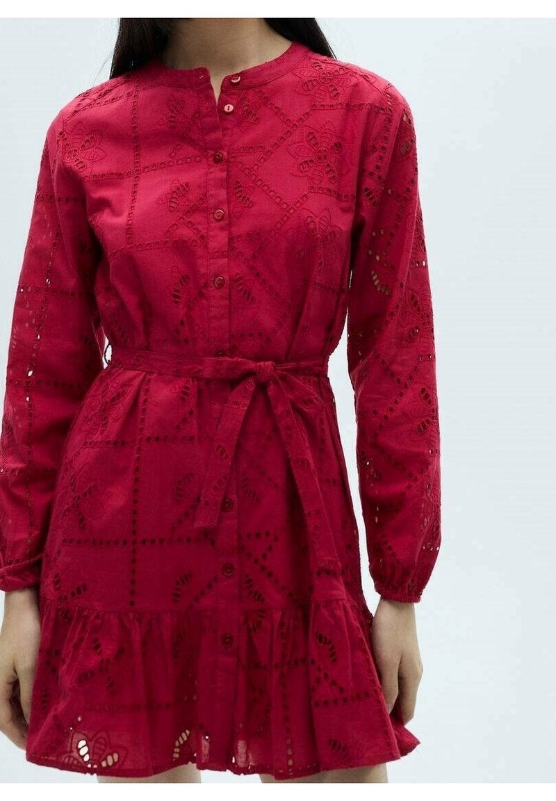 Femme portant une robe rouge à manches longues avec des motifs floraux ajourés, des boutons sur le devant, une ceinture nouée à la taille et un ourlet à volants.