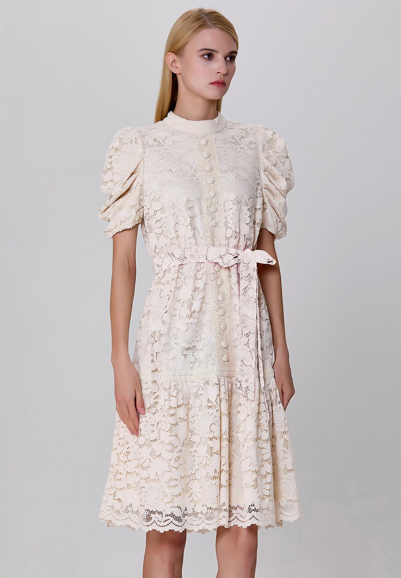 Robe en dentelle de couleur crème clair avec des manches bouffantes, un devant à boutons et une ceinture à nouer. Présente des motifs floraux et un ourlet festonné.
