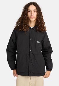 Jong persoon met lang krullend haar, gekleed in een zwarte jas met knopen en elastische manchetten, en beige broek, tegen een effen achtergrond.