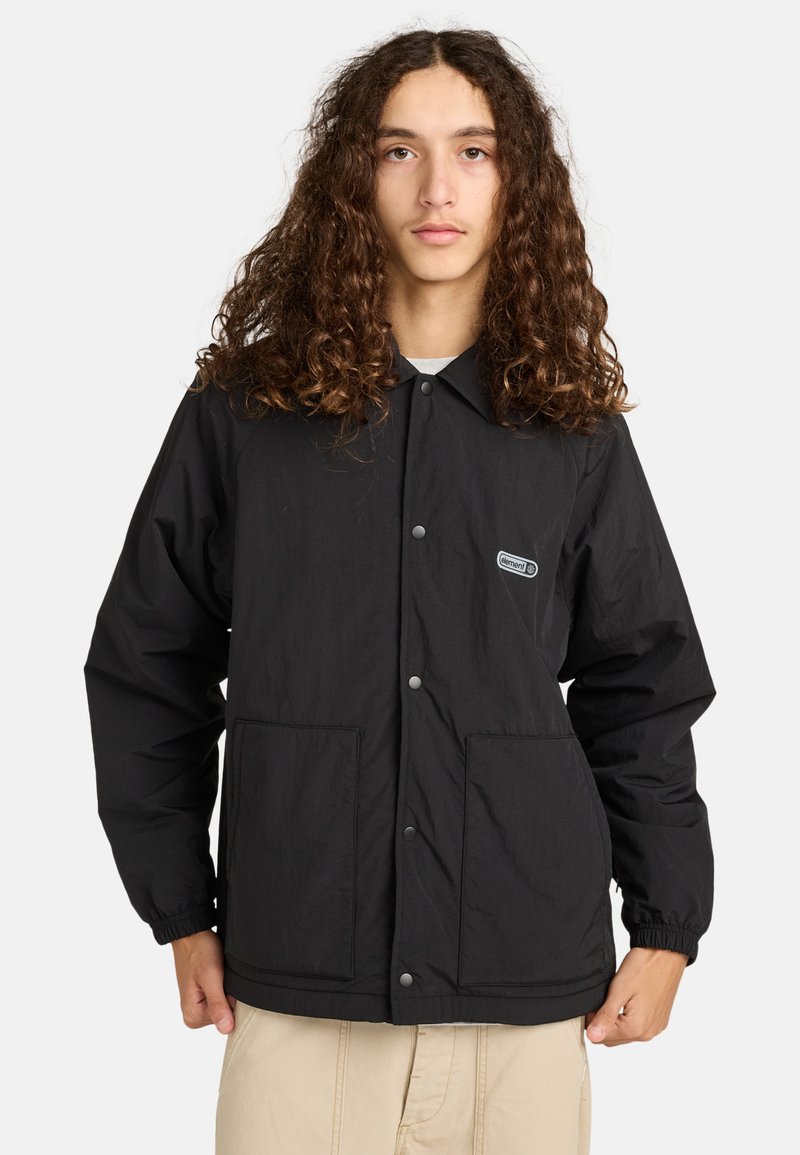 Jong persoon met lang krullend haar, gekleed in een zwarte jas met knopen en elastische manchetten, en beige broek, tegen een effen achtergrond.