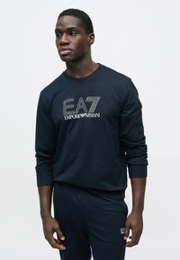 Tengerészkék hosszú ujjú pulóver "EA7 Emporio Armani" logóval. Bordázott mandzsettákkal és kerek nyakkivágással rendelkezik, puha pamut anyagból készült.