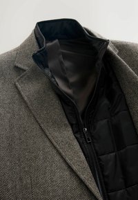 Sacou maro cu model în chevron, dotat cu căptușeală neagră căptușită și cu fermoar. Prezintă un guler și reveruri ajustate, evidențiind un material texturat.