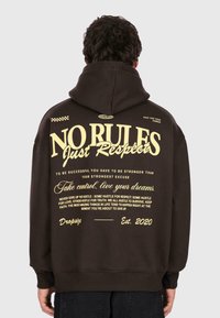 Personne portant un sweat à capuche noir avec du texte jaune au dos : "PAS DE RÈGLES Juste du Respect," phrases motivantes, et "Dropsiz Est. 2020."